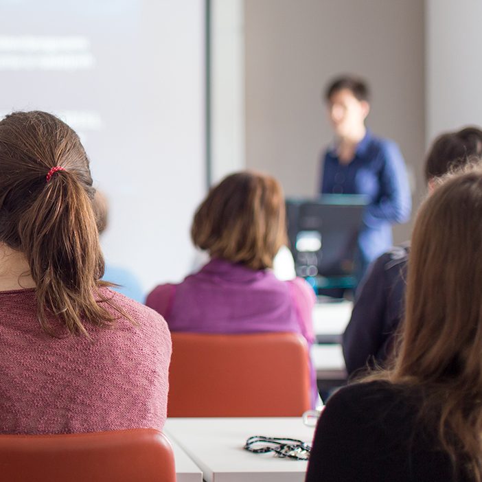 Female speaker giving presentation in lecture hall at university workshop. Rear view of unrecognized participants listening to lecture and making notes. Scientific conference event.